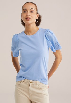 Žena v svetlomodrom krátkom rukáve s háčkovanými rukávmi a béžovými nohavicami stojaca so založenými rukami za chrbtom.