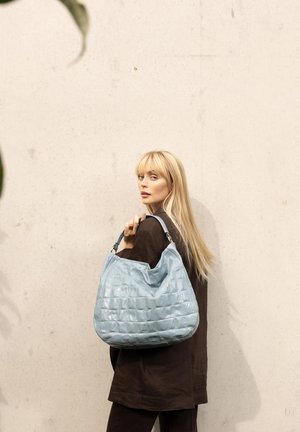 Blonde woman in dark outfit holding a large light blue textured handbag, standing against a plain beige wall.
