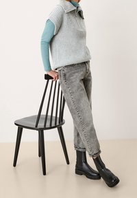 Stickad grå väst med krage över en teal långärmad skjorta, kombinerad med blekta grå jeans och svarta ankelstövlar, lutad mot en svart stol.