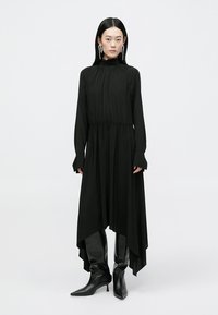 Μαύρο φόρεμα με μακριά μανίκια, ψηλό λαιμό και ζωνάκι στη μέση, με ασύμμετρο τελείωμα και ρευστή σιλουέτα, φορεμένο με μπότες μέχρι το γόνατο.