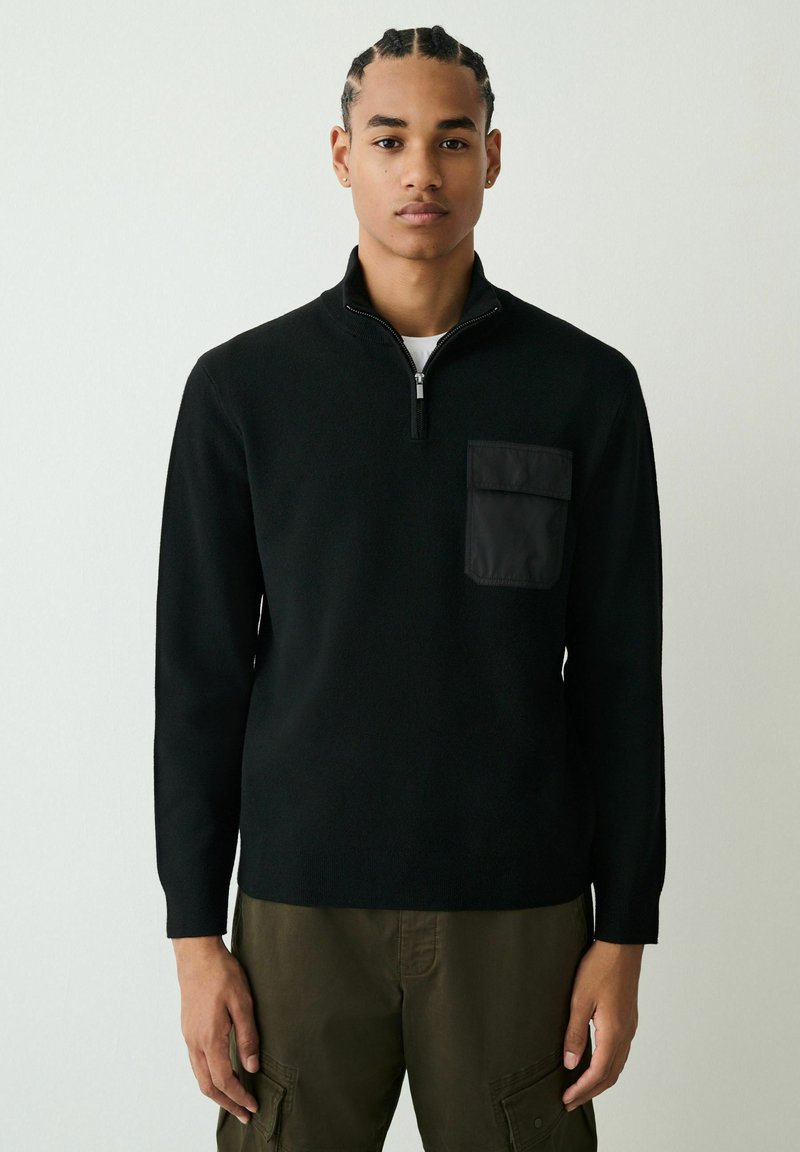 Crni flis pulover s četvrtinskim patentnim zatvaračem i džepom na prsima. Ima duge rukave i mekanu teksturu, upareno s cargo stilom zelenih hlača.