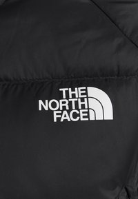 Giubbotto piumino nero con un design trapuntato, caratterizzato da un evidente logo bianco "The North Face" nella zona del petto. Texture liscia.