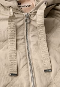 Beige gewatteerde jas met een ritssluiting, gestructureerde capuchon en gelabelde koordjes. Bevat zilverkleurige hardware en een gladde stofafwerking.