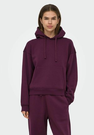 Jeune femme portant un sweat à capuche violet foncé et un pantalon de jogging assorti, debout avec une main dans la poche devant un fond clair uni.