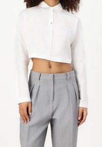 Camicia bianca a maniche lunghe con colletto a punta e chiusura con bottoni, abbinata a pantaloni grigi chiari con pieghe su misura.