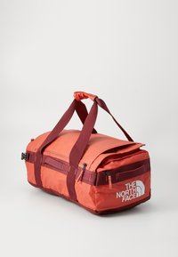Red and maroon duffel bag with two handles, zippered compartments, and "The North Face" logo on the side, set against a plain light background.