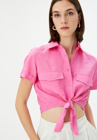 Camiseta corta de manga corta en color rosa con diseño de nudo en la parte delantera, que presenta un cuello tipo solapa y dos bolsillos en el pecho. Tela suave con una textura ligeramente arrugada.