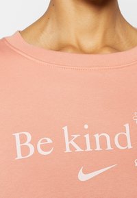 Ljust rosa sweatshirt med rund halsringning, som har texten "Var snäll" och en vit Nike swoosh-logotyp. Mjuk textur och enkel design.