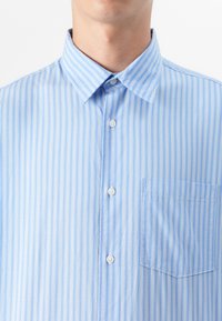 Primer plano del torso superior de un hombre que lleva una camisa de botones de rayas verticales azul claro con cuello y un bolsillo en el pecho.