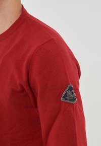 Maglione rosso lavorato a maglia con scollatura rotonda e finitura testurizzata. Presenta una toppa triangolare con il nome del marchio sulla manica sinistra.