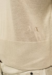 Μπεζ πλεκτό πουλόβερ με ριμπ τελείωμα και διακριτικό κεντητό λογότυπο "A|X" στην κάτω πλευρά πάνω από μπεζ παντελόνια με σχέδιο.