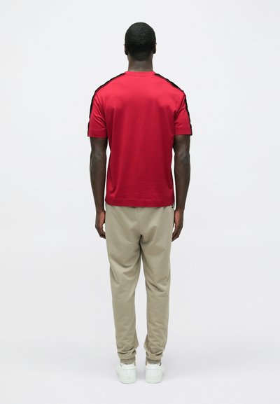 Rdeča majica s kratkimi rokavi s črnimi črtami na ramenih, v kombinaciji z bež trenirkami in belimi supergami, pogled od zadaj.