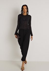 Top de malla negro de manga larga con puntos plateados, combinado con pantalones negros holgados y sandalias de tacón beige. Sosteniendo un pequeño bolso de mano marrón.