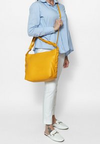 Borsa a tracolla gialla in pelle con manico intrecciato, forma rettangolare, texture liscia e zip superiore, indossata da una persona con abbigliamento chiaro.