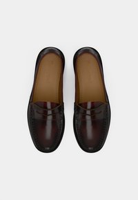 Brūni ādas loaferi ar spīdīgu apdari, kas izceļas ar dekoratīvu šuvumu un noapaļotu purngalu. Izmīkstināts iekšpuse, kas ir viegli brūnā krāsā.