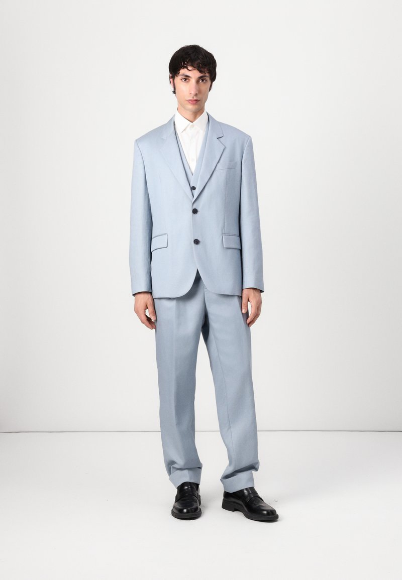 Traje azul claro con chaqueta a medida y pantalones a juego. Presenta botones negros, un bolsillo en el pecho y una camisa blanca debajo.