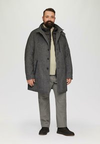 Homme avec une barbe portant un manteau gris à couches, un pull beige, un pantalon gris et des chaussures noires se tenant devant un fond uni.