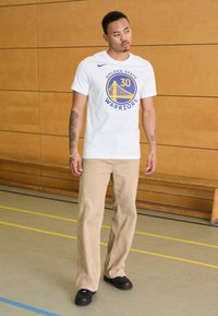 Hvid T-shirt med "Golden State Warriors" logo i blå, beige bukser og sorte sko. Glat stof, afslappet pasform og synlige grafikker.