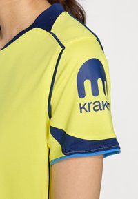Žuta atletska majica s tamnoplavim akcentima, s okruglim izrezom, detaljima šivanja i velikim logotipom na rukavu.