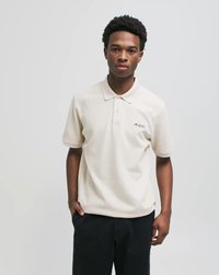 Mladi moški s kratkimi kodrastimi lasmi nosi kremno polo majico z majhnim logotipom in črne hlače, stoji ob enobarvni svetli podlagi.