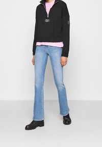 Svart dragkedja pullover med en framsidan logotyp över en rosa skjorta, ihopparad med ljusblå utsvängda jeans och svarta ankelskor.