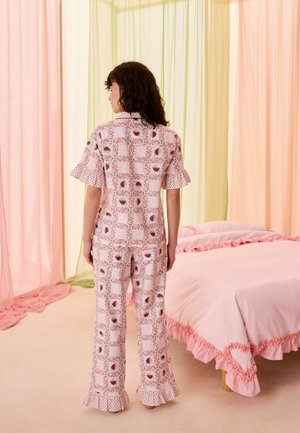 Donna in pigiama rosa a fantasia con maniche e pantaloni arricciati, in piedi accanto a un letto con biancheria da letto rosa arricciata e tende pastello.