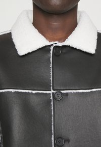 Giacca di pelle nera con colletto e dettagli in pelliccia bianca, chiusa con grandi bottoni neri su una carnagione scura.