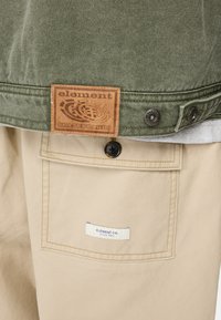Chaqueta de lona verde con una etiqueta de cuero marrón. Pantalones beige con un bolsillo y etiqueta azul. Botón de metal visible en la chaqueta.
