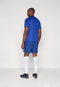 Blå atletisk skjorte med logomønster, parret med matchende shorts. Modellen har hvide knæstrømper og sorte sportsko på. Minimalistisk baggrund.