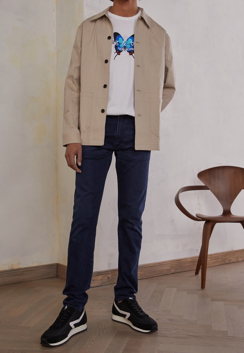 Beige knäppt jacka över en vit t-shirt med en blå fjärils grafik, matchat med mörk denim jeans och svarta sneakers med vita detaljer.