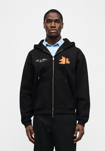 Νεαρός άνδρας φοράει μαύρο φούτερ με φερμουάρ και πορτοκαλί γραφικό με φλόγα και κείμενο στο αριστερό στήθος, ανοιχτό μπλε πουκάμισο με γιακά από κάτω, στέκεται ουδέτερα.