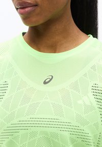 Κοντινό πλάνο ενός ατόμου που φοράει ανοιχτοπράσινο αθλητικό πουκάμισο με τρύπες και λογότυπο Asics στο στήθος.