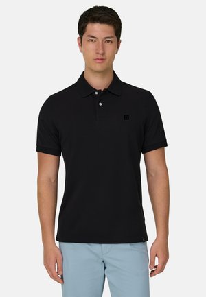 Joven con polo negro de manga corta y pantalones azul claro, posando frente a un fondo gris claro liso.