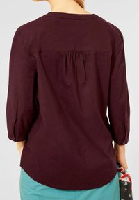 Femme portant un chemisier à manches longues violet foncé avec un détail froncé dans le dos, tenant un tissu à motifs dans la main droite, et portant un pantalon bleu sarcelle.