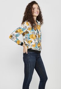 Giacca bomber floreale con motivi di ibisco gialli e blu su sfondo bianco, con polsini e orlo a coste neri. Indossata con jeans scuri.