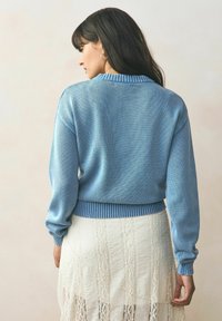Pulover bleu tricotat cu mansete și tiv în dantelă, purtat peste o fustă albă din dantelă, evidențiind o textură moale și un croi relaxat.