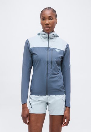 Kobieta w dwukolorowej, niebieskiej kurtce na zamek z kapturem i jasnoniebieskich szortach stoi na białym tle.