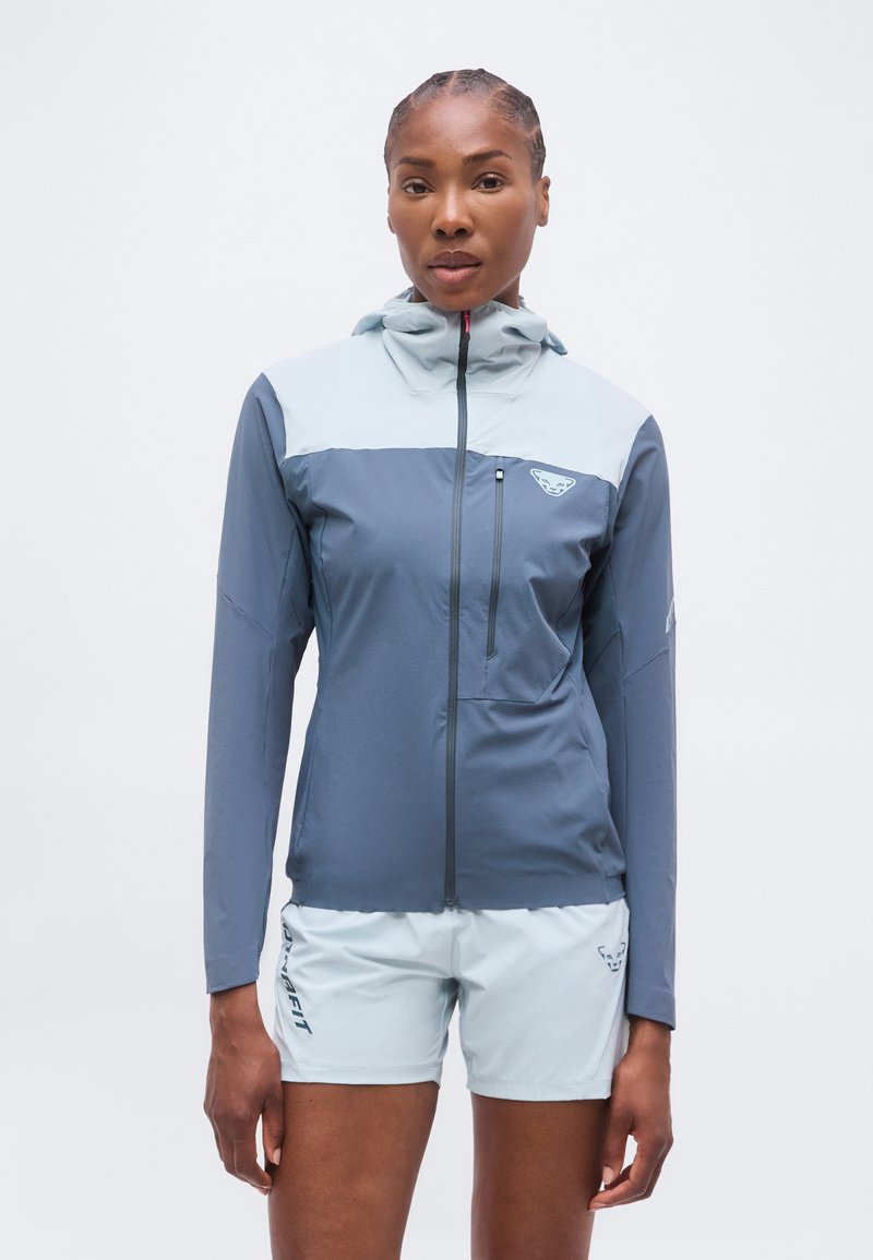 Γυναίκα με δίχρωμο μπλε φερμουάρ-φούτερ με κουκούλα και ανοιχτό μπλε σορτς, στέκεται μπροστά σε ένα λευκό φόντο.