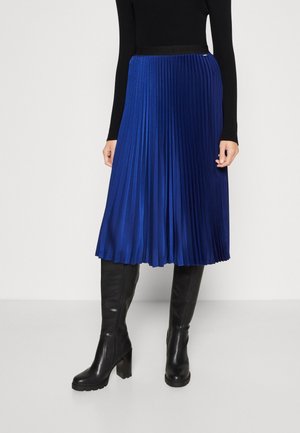 Femme portant une jupe midi plissée bleue, un haut noir à manches longues et des bottes noires à talons jusqu'aux genoux, debout devant un fond uni.
