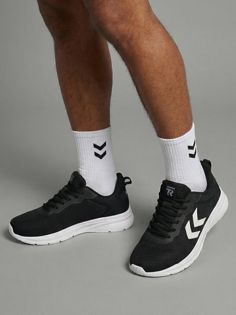 Persona che indossa sneakers nere in mesh con suole bianche e calzini bianchi alti con loghi a chevron neri, in piedi su pavimento grigio.