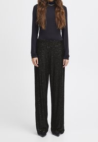 Pantalon noir en sequins avec une coupe droite, présentant une surface texturée et des accents réfléchissants. Associé à un haut noir à manches longues.
