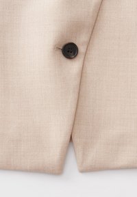 Gros plan d’un tissu beige avec un seul bouton foncé cousu à côté d’une couture verticale sur un vêtement.