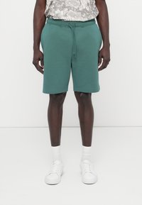 Πράσινα βαμβακερά σορτς με ελαστική μέση και κορδόνι, διαθέτουν πλαϊνές τσέπες και ίσια γραμμή, συνδυασμένα με λευκά αθλητικά παπούτσια.