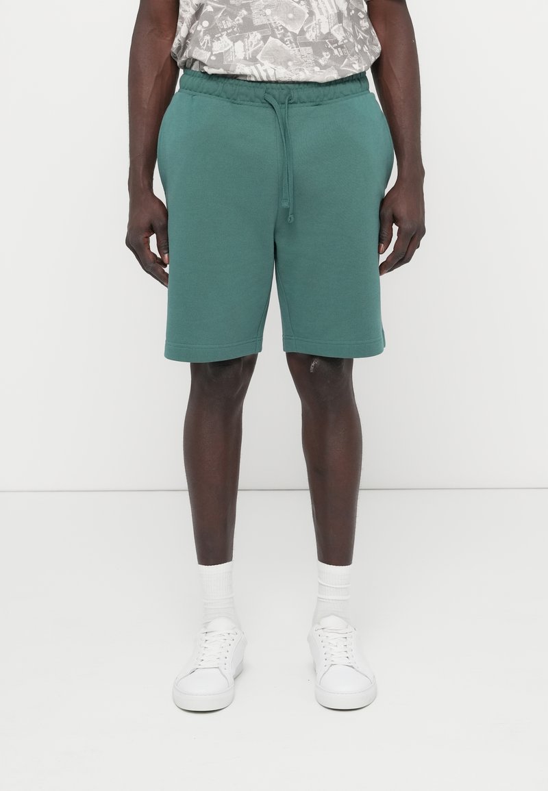 Πράσινα βαμβακερά σορτς με ελαστική μέση και κορδόνι, διαθέτουν πλαϊνές τσέπες και ίσια γραμμή, συνδυασμένα με λευκά αθλητικά παπούτσια.