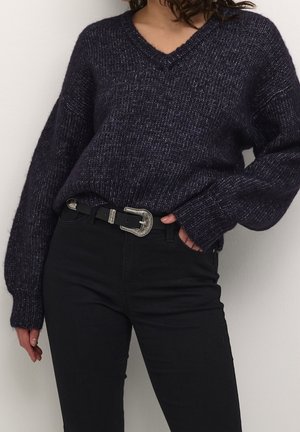 Žena nosící tmavě fialový pletený svetr s výstřihem do V, zastrčený do černých kalhot s vysokým pasem, doplněných černým páskem se stříbrnou přezkou.