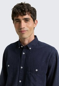 Herren Dunkelblaues Button-Down-Hemd, strukturierter Stoff mit Sprenkeln, Kragen, zwei Taschen und weißen Knöpfen.