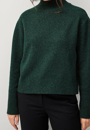 Femme portant un pull à col roulé côtelé vert foncé et un pantalon noir, les mains détendues le long du corps.