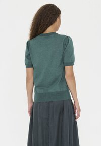 Kratke sleeves, temno zelena pletena majica z ribastim robom in napihnjenimi rameni. Mehka tekstura, lahkoten material, minimalen dizajn.