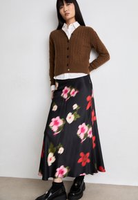 Brun kabelstrikket cardigan, hvit skjorte med knapper, svart satin maxiskjørt med blomstmønster, og svarte platåsko.