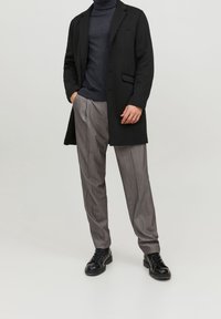 Schwarzer Mantel über einem dunklen Rollkragenpullover, kombiniert mit grauen gestreiften Hosen. Schwarze Schnürschuhe runden das Outfit ab. Schlichte Hintergrund.